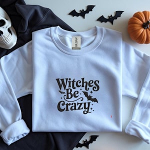 Könnte beinhalten: Weißes Sweatshirt mit Rundhalsausschnitt und dem Text "Witches Be Crazy" in Schwarz. Das Bild zeigt einen Totenkopf, einen kleinen orangefarbenen Kürbis und schwarze Fledermaus-Ausschnitte, die ein Halloween-Thema erzeugen.