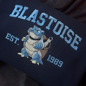 Könnte beinhalten: Marineblaues Sweatshirt mit hellblau gesticktem Schriftzug "BLASTOISE" über einer gestickten Cartoon-Figur von Blastoise. Der Text "EST. 1989" ist ebenfalls auf dem Sweatshirt gestickt.