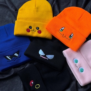 May include: A collection of colorful knit beanies, each embroidered with a different cartoon character face. The beanies are in various colors, including yellow, orange, blue, black, and pink. The beanies are made of a soft, knitted material.