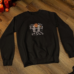 May include: Black sweatshirt with a Halloween design. The graphic shows two skeletons dancing in front of a pumpkin. The design uses white, orange, and black. The sweatshirt has a crew neck and long sleeves.