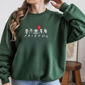 May include: Dark green sweatshirt with a graphic of horror movie characters in the style of the TV show "Friends." The graphic includes characters like Pennywise, Ghostface, and Michael Myers, with a red balloon. The word "FRIENDS" is below the characters.