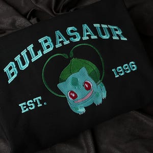 Op de afbeelding: Zwart sweatshirt met turquoise borduursel. Het woord "BULBASAUR" staat bovenaan in een boog, met "1996" rechts en "EST." links. In het midden staat een turquoise Bulbasaur-personage met rode ogen en een lachend gezicht.