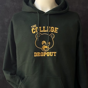 May include: Dark green hoodie with a kangaroo pocket and drawstring hood. The front features gold embroidered text and a bear graphic that reads "THE COLLEGE DROPOUT". The sweatshirt is displayed on a mannequin.