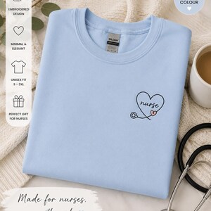 May include: A light blue crew neck t-shirt featuring a small embroidered design on the left chest. The embroidery consists of a black heart outline with the word nurse written inside in cursive, accompanied by a stethoscope and a small pink heart.