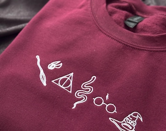 Puede incluir: Sudadera burdeos con bordado blanco de un diseño lineal de una varita, una snitch dorada, un triángulo con un círculo, una serpiente, gafas y un sombrero seleccionador. La sudadera está doblada sobre una superficie oscura y texturizada.