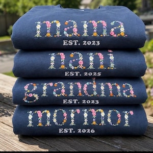 Peut inclure: Une pile de quatre sweat-shirts bleu marine à col rond avec des lettres à motifs floraux. Les vêtements portent les mots mama, nani, grandma et mormor, chacun accompagné du texte EST. suivi d'une année, soit 2023, 2025, 2023 et 2026.