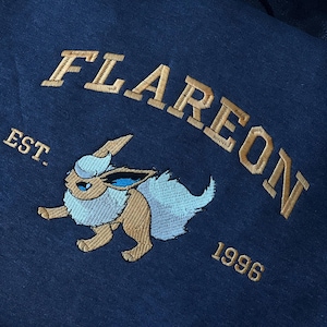 May include: Navy blue hooded sweatshirt with gold embroidered text and a cartoon character. The word "FLAREON" arches across the top, with "EST. 1996" below. The character is brown, white, and blue.