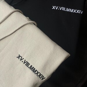 May include: Two hoodies, one black and one beige, are shown in a close-up shot. Both have white embroidered text: XV.VIII.MMXXIV. The beige hoodie has a drawstring. The black hoodie is partially visible, with the beige hoodie in the foreground.