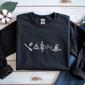 May include: Black crew neck sweatshirt featuring white line drawings of a broom, lightning bolt, triangle, snake, glasses, and sorting hat. The sweatshirt is shown with blue jeans.