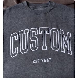 Peut inclure: Un sweat-shirt gris foncé avec le mot "CUSTOM" en grandes lettres blanches sur la poitrine. En dessous, le texte "EST. YEAR" est écrit en plus petits caractères blancs. Le sweat-shirt a un col rond.