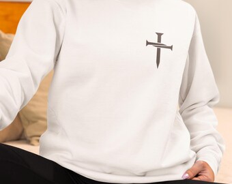 Puede incluir: Una sudadera blanca con un diseño de cruz y clavo gris oscuro en la parte superior izquierda del pecho. La parte superior de manga larga está hecha de un material suave. La persona lleva leggings negros.