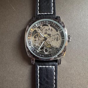 May include: A square-faced, skeleton watch with a black leather band and white stitching. The watch face displays Roman numerals and intricate internal gears. The watch has a silver-colored bezel and crown.