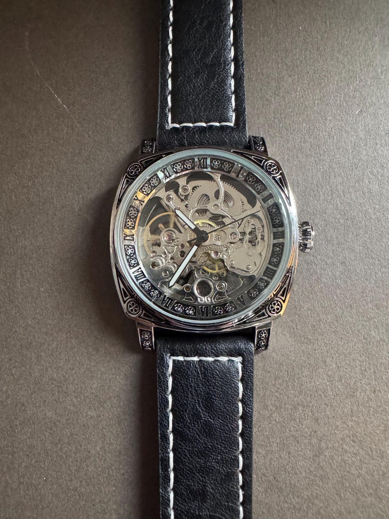 May include: A square-faced, skeleton watch with a black leather band and white stitching. The watch face displays Roman numerals and intricate internal gears. The watch has a silver-colored bezel and crown.