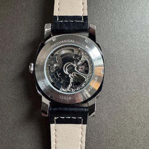 May include: A stainless steel mechanical watch with a black leather strap and a cream-colored underside. The watch's back is transparent, revealing the intricate movement. The words "MECHANICAL" and "STAINLESS STEEL" are visible.