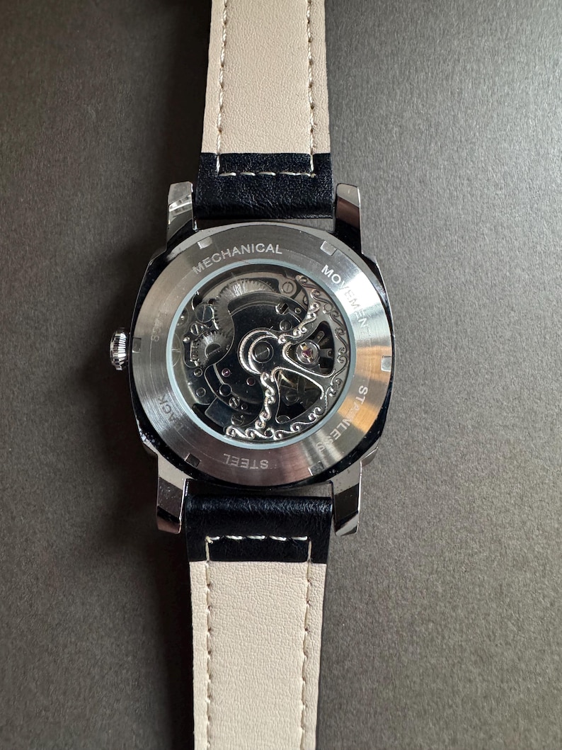 May include: A stainless steel mechanical watch with a black leather strap and a cream-colored underside. The watch's back is transparent, revealing the intricate movement. The words "MECHANICAL" and "STAINLESS STEEL" are visible.