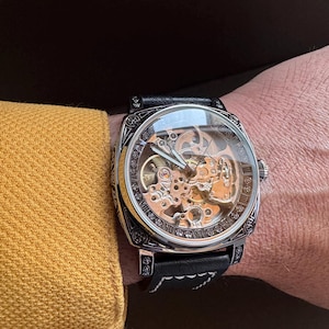 May include: A skeleton watch with a black leather band. The watch has a silver-colored case with intricate detailing and a clear face, revealing the inner workings. The watch is worn on a wrist next to a yellow sleeve.