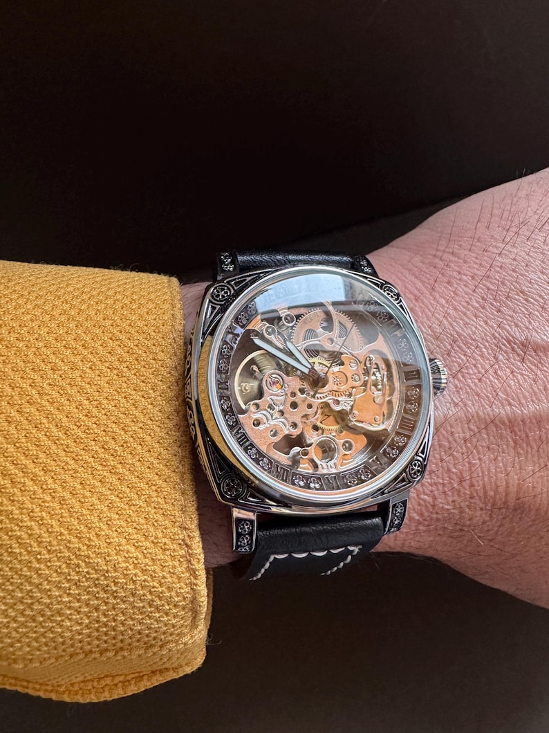 May include: A skeleton watch with a black leather band. The watch has a silver-colored case with intricate detailing and a clear face, revealing the inner workings. The watch is worn on a wrist next to a yellow sleeve.