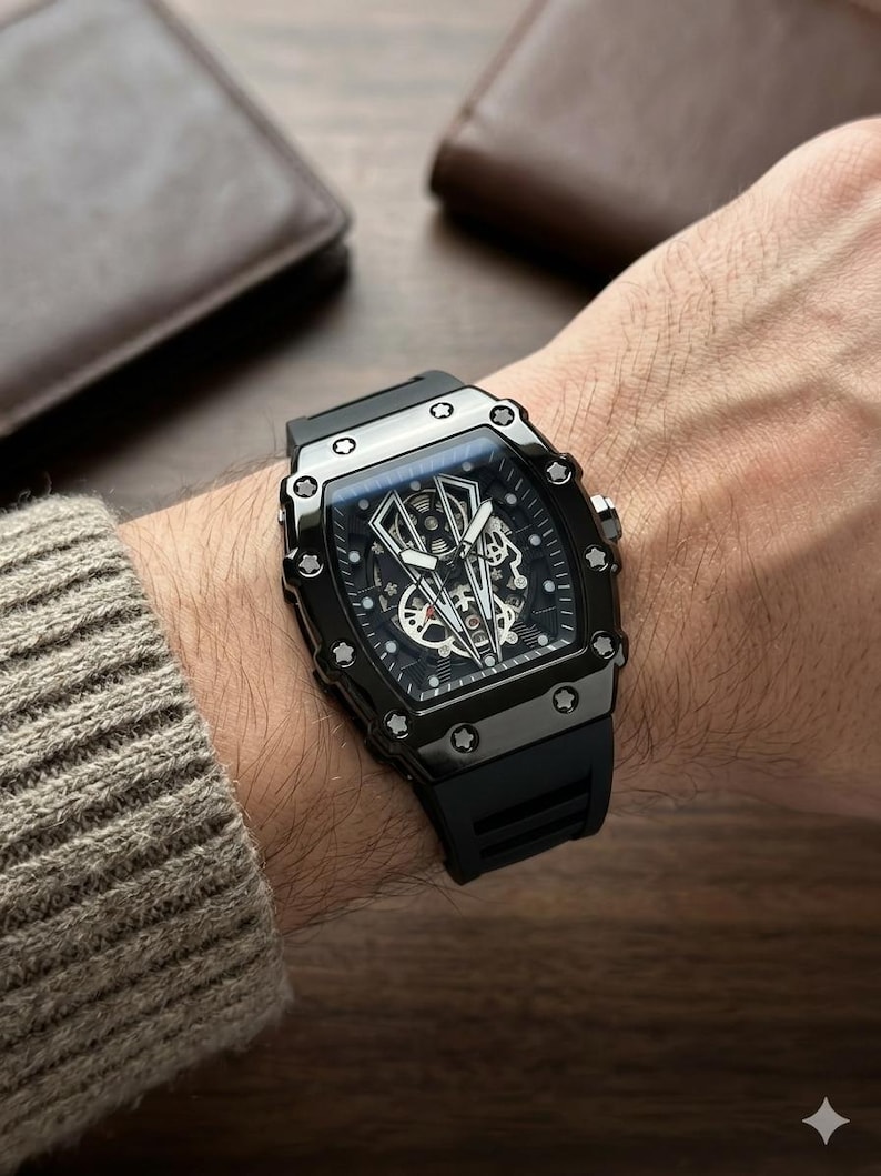 May include: A black wristwatch with a rectangular face and a black rubber strap. The watch has a detailed dial with visible gears and a dark, metallic case. The watch is on a wrist, with a brown leather wallet in the background.
