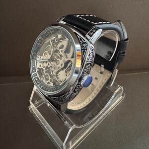 May include: A silver-toned skeleton watch with a black leather band and white stitching. The watch features intricate internal mechanisms visible through the face and decorative detailing around the bezel. A blue stone accents the side of the watch.