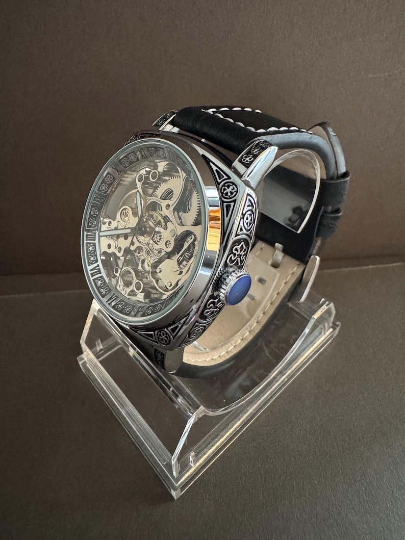 May include: A silver-toned skeleton watch with a black leather band and white stitching. The watch features intricate internal mechanisms visible through the face and decorative detailing around the bezel. A blue stone accents the side of the watch.