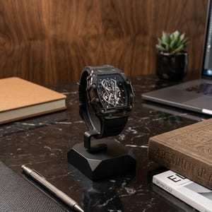 May include: A black wristwatch with a skeleton dial is displayed on a black stand. The watch has a black strap and a complex internal mechanism visible through the face. The watch is on a black marble surface.
