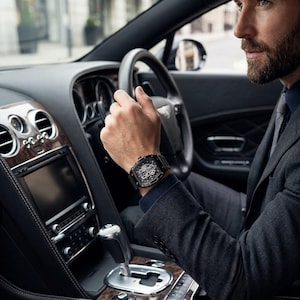 May include: A black wristwatch with a detailed face is worn on a person's wrist. The watch is set against the backdrop of a luxury car's interior, with wood and leather detailing. The person is wearing a dark suit and is driving.