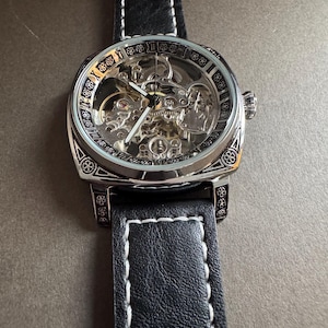 May include: A silver-toned skeleton watch with a black leather band. The watch face reveals the intricate inner workings, with Roman numerals around the edge. The black band has white stitching.