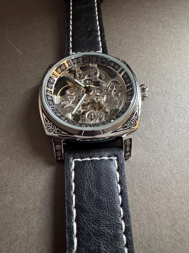May include: A silver-toned skeleton watch with a black leather band. The watch face reveals the intricate inner workings, with Roman numerals around the edge. The black band has white stitching.