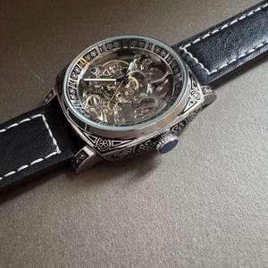 May include: A silver-toned skeleton watch with a black leather band and white stitching. The watch face reveals the intricate inner workings. The watch has a blue crown and is set against a neutral background.