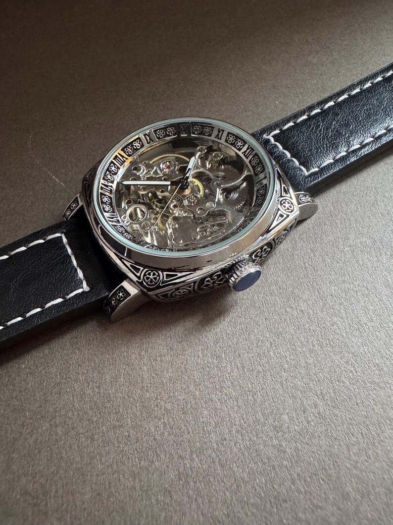 May include: A silver-toned skeleton watch with a black leather band and white stitching. The watch face reveals the intricate inner workings. The watch has a blue crown and is set against a neutral background.