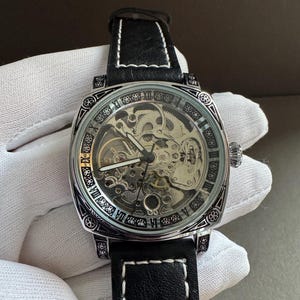 May include: A silver-toned, skeleton-dial wristwatch with a black leather band and white stitching. The watch features Roman numeral hour markers and intricate internal gears visible through the clear face. The watch has a square face.