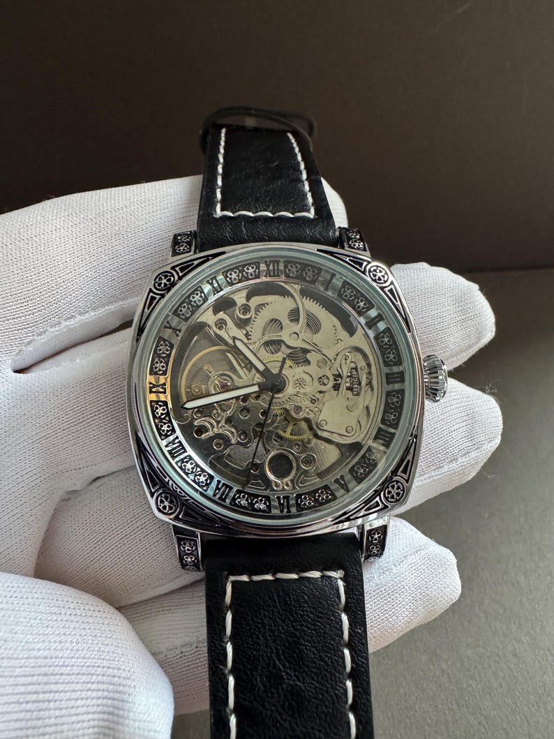 May include: A silver-toned, skeleton-dial wristwatch with a black leather band and white stitching. The watch features Roman numeral hour markers and intricate internal gears visible through the clear face. The watch has a square face.