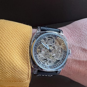 May include: A skeleton watch with a black leather band. The watch face is clear, revealing the intricate inner workings. The watch has a silver-colored bezel with decorative patterns and Roman numerals. The watch is on a wrist.