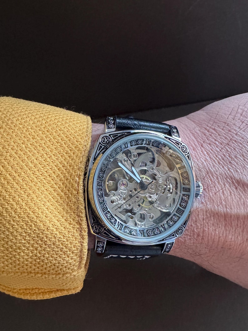 May include: A skeleton watch with a black leather band. The watch face is clear, revealing the intricate inner workings. The watch has a silver-colored bezel with decorative patterns and Roman numerals. The watch is on a wrist.
