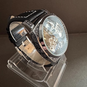 May include: A silver and black skeleton watch with a clear face, showcasing the intricate inner workings. The watch has a black leather band with white stitching and is displayed on a clear acrylic stand. The watch case has decorative patterns.
