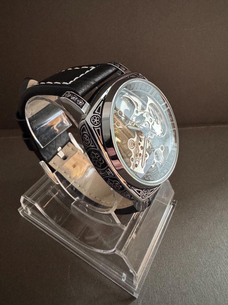 May include: A silver and black skeleton watch with a clear face, showcasing the intricate inner workings. The watch has a black leather band with white stitching and is displayed on a clear acrylic stand. The watch case has decorative patterns.