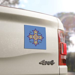 May include: A blue square magnet featuring a yellow and blue cross design with stylized text. The cross has intricate patterns and is centered on the magnet. The magnet is attached to the side of a white vehicle.