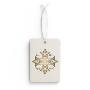 May include: A rectangular, off-white air freshener with a gold-colored, ornate cross design. The cross features intricate patterns and text. A white string is attached for hanging. The design is centered on the air freshener.