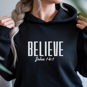 Puede incluir: Sudadera con capucha negra con la palabra "BELIEVE" en letras blancas, con "John 14:1" debajo. La sudadera tiene una capucha con cordón y un bolsillo delantero. El color es negro.