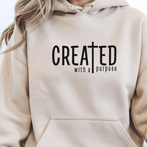 Puede incluir: Sudadera con capucha color arena con bolsillo delantero y capucha. El texto negro en la parte delantera dice "CREATED with a purpose" con una cruz incorporada en la palabra "CREATED". La sudadera es de color neutro y tiene un ajuste relajado.