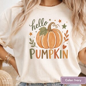 May include: An ivory t-shirt featuring a graphic print of an orange pumpkin with a green leaf. The text "hello PUMPKIN" is printed in a mix of green and orange colours. The design includes small green and orange stars, leaves, and a heart.
