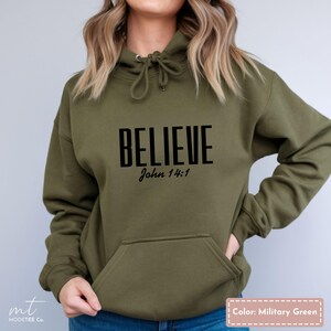 Puede incluir: Sudadera con capucha verde oliva con la palabra "BELIEVE" en letras negras y en negrita, con "John 14:1" debajo. La sudadera tiene un bolsillo delantero y una capucha con cordón. El color se describe como "Verde militar".