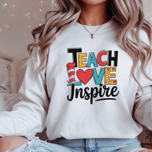 Puede incluir: Sudadera blanca con las palabras "Teach Love Inspire" en un diseño colorido. El diseño incluye un corazón rojo, una "V" azul y un sombrero a rayas rojas y blancas. Prenda casual.