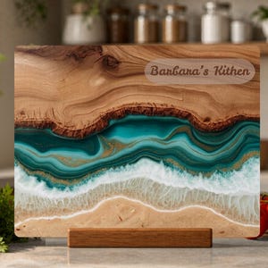 Op de afbeelding: Houten snijplank met een uniek ontwerp met een strandscène met turquoise water en zandstranden. De plank heeft een houten voet en de tekst "Barbara's Kithen". De plank is gemaakt van hout en hars.