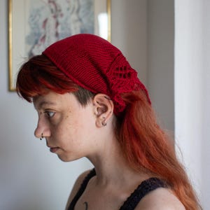 May include: A vibrant red knitted headscarf featuring a textured stitch pattern along the edges. The accessory is crafted from yarn and designed to be worn over the hair, covering the crown while leaving the sides and back of the head exposed.