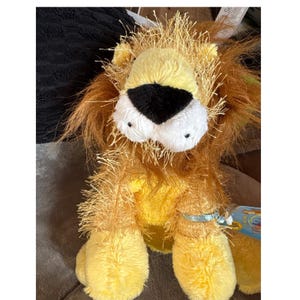 May include: A plush lion toy with a yellow body, a yellow and black nose, and white eyes. The lion has a shaggy, brown mane and a tag attached to its leg. The toy is designed to look like a friendly lion.