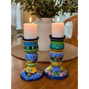 May include: Two hand-painted ceramic candle holders with lit white candles. The candle holders are blue, green, yellow, and pink with floral and geometric designs. The candles are white and cylindrical, and the flames are orange.