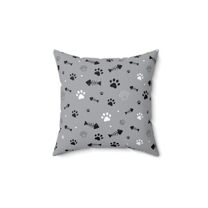 Op de afbeelding: Vierkant kussen met een grijze achtergrond en een herhalend patroon van zwarte en witte pootafdrukken en visgraten. Een decoratief item voor in huis.