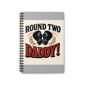 May include: Spiral-bound notebook with a gray cover and the words "ROUND TWO DADDY!" in black and red. The design includes black boxing gloves and a laurel wreath on a cream background.