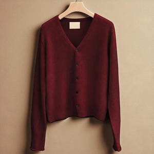 Puede incluir: Un cárdigan burdeos con cuello en V, mangas largas y cierre de botones. El suéter está colgado en una percha de madera. El suéter tiene dobladillo y puños acanalados. El fondo es neutro.